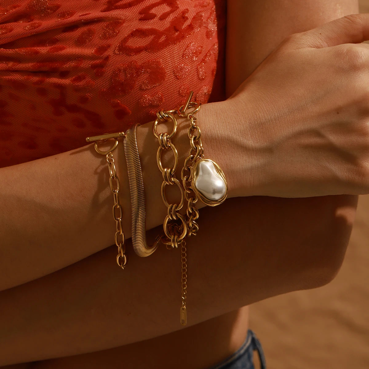 Gold stacked chain bracelet with pearl charm on wrist against red textured top