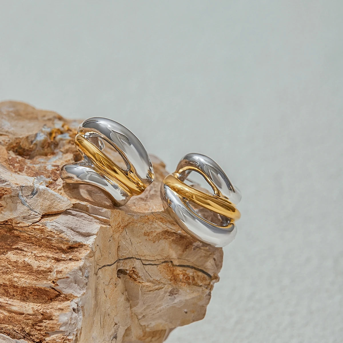 Luxury stud earrings with intertwined silver and gold bands displayed on natural stone