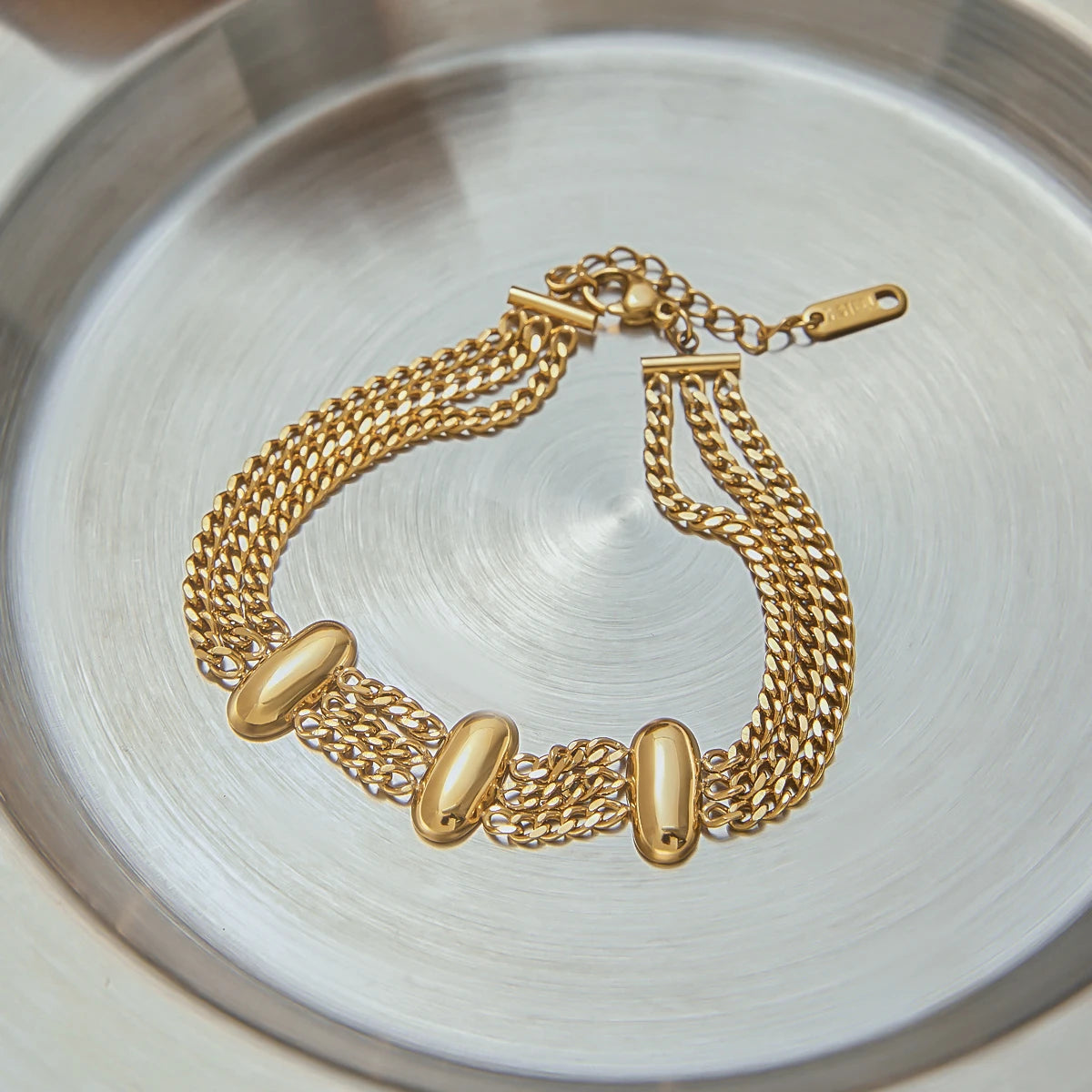 Fashionable gold bracelet with triple chain design and oval accents displayed on silver tray