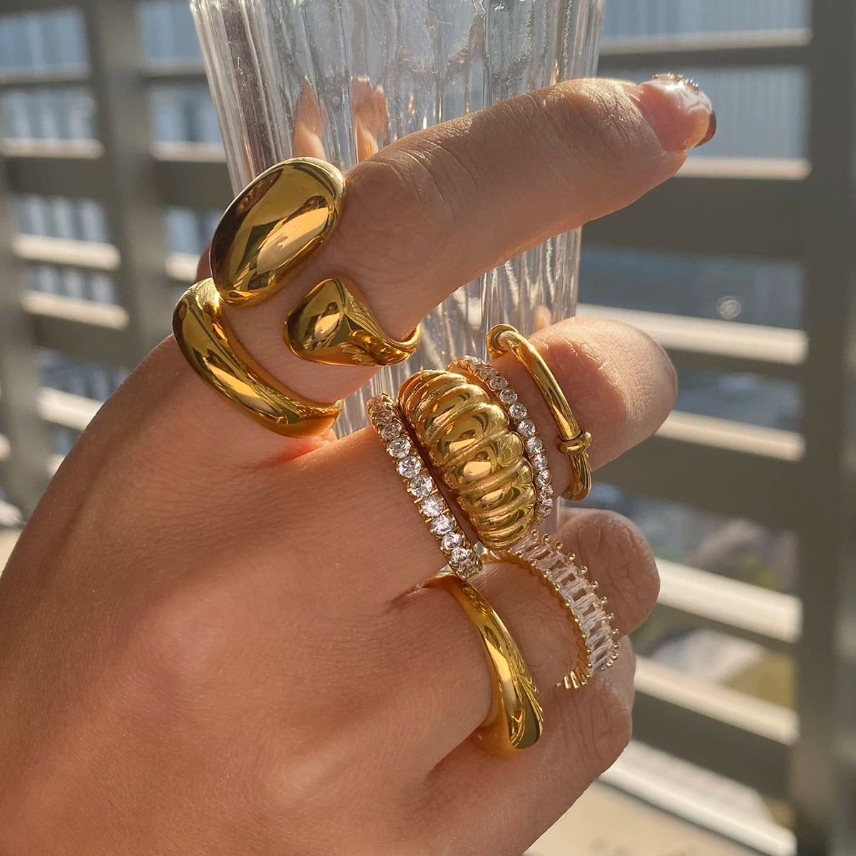 Hand wearing shiny gold and diamond Fashionable Stackable Rings with various designs near a glass cup