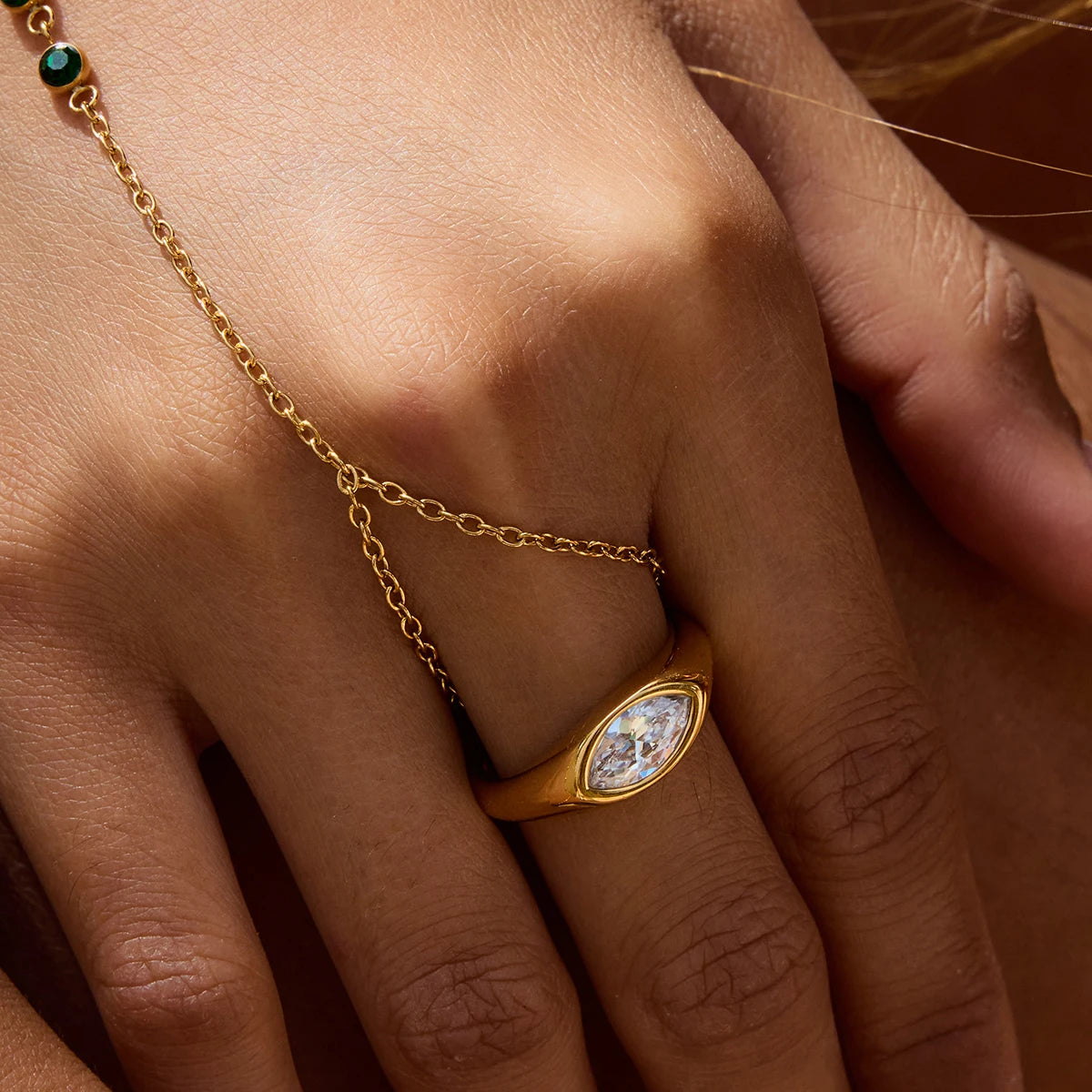 Gold geometric statement ring with marquise-cut clear gemstone worn on finger with gold chain bracelet
