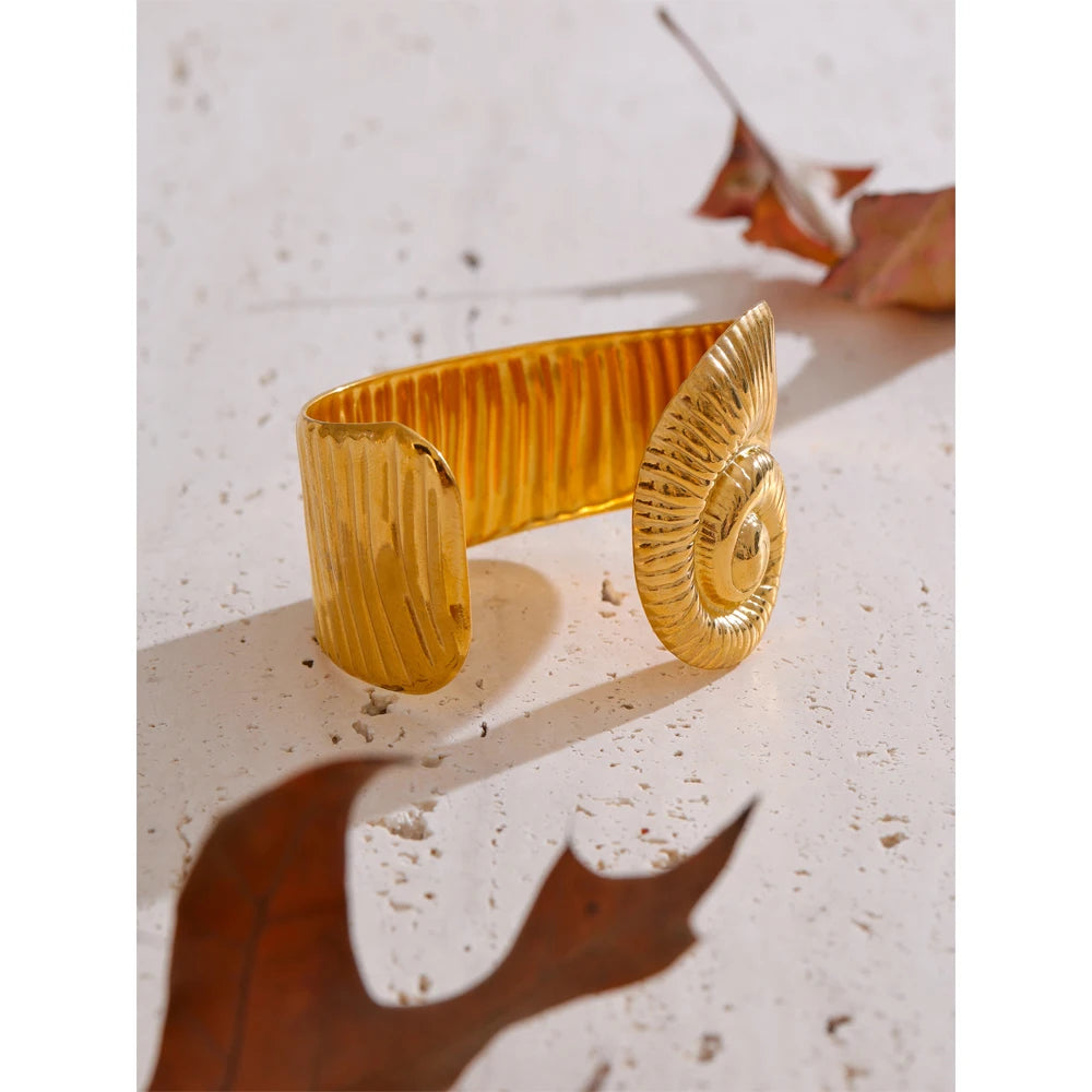Gold waterproof open bangle with a spiral textured design displayed on a light surface with dried leaves